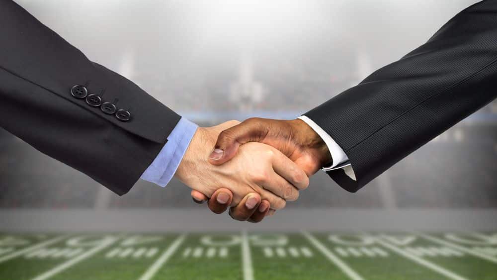 men in suits doing a handshake in front of a football field.