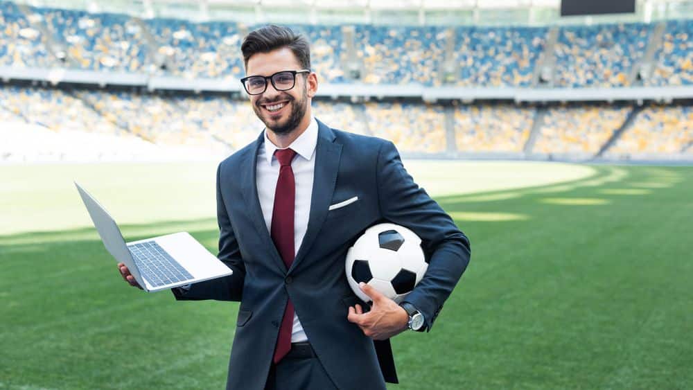 suited man holding a ball and a laptop standing on a football field.