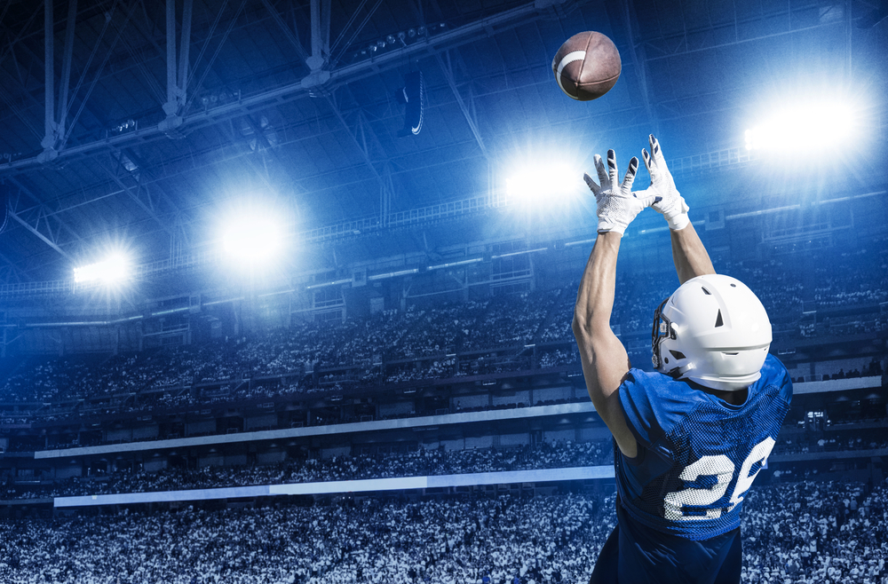 A player in a blue jersey with white helmet reaches up to catch a football in front of a massive crowd.