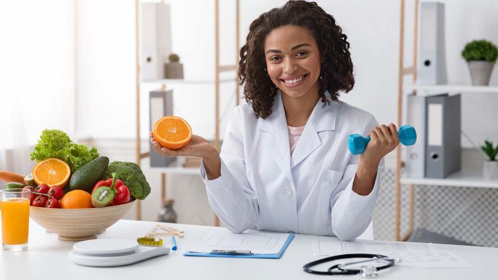 Smiling sports dietitian in uniform holding an orange and a weight