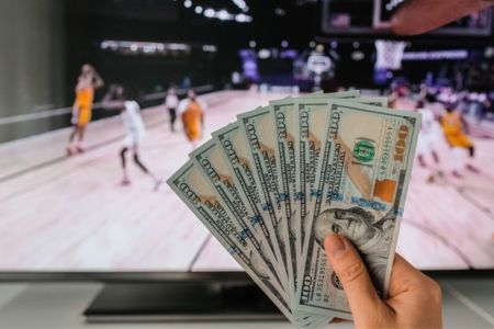 Hand with hundred dollar bills and basketball game on tv in background