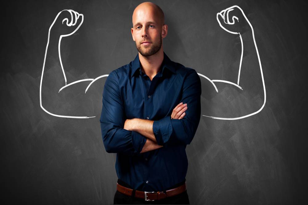 Sports Account manager stands in front of a blackboard with flexing arms draw behind him in chalk.