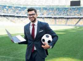 A sports marketer holding a laptop and soccer ball in a soccer stadium.