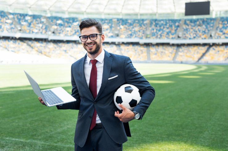 A sports marketer holding a laptop and soccer ball in a soccer stadium.