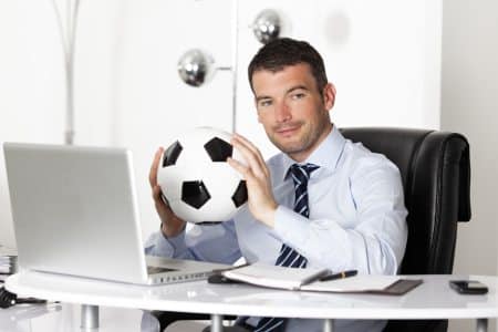 A sports tourism event planner holding a soccer ball while sitting at a desk with a laptop on it and looking at the camera.