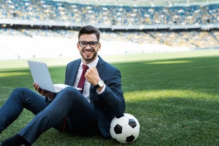 A sports tourism manager sitting in a soccer stadium while holding a laptop and smiling at the camera.