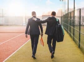 two men carrying tennis gear across the court.