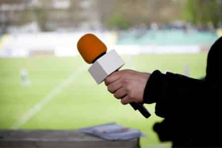 person holding a microphone with a football match in the background.